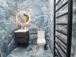 A bathroom with a marble wall, a round mirror, a sink, a toilet, and a black metal rack. - Cottage by the River (Burnley)