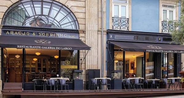 Uma área de refeições ao ar livre com mesas e cadeiras, fachada de vidro e placa com a inscrição 'Araujo & Sobrinho, Restaurante Galeria do Largo'.