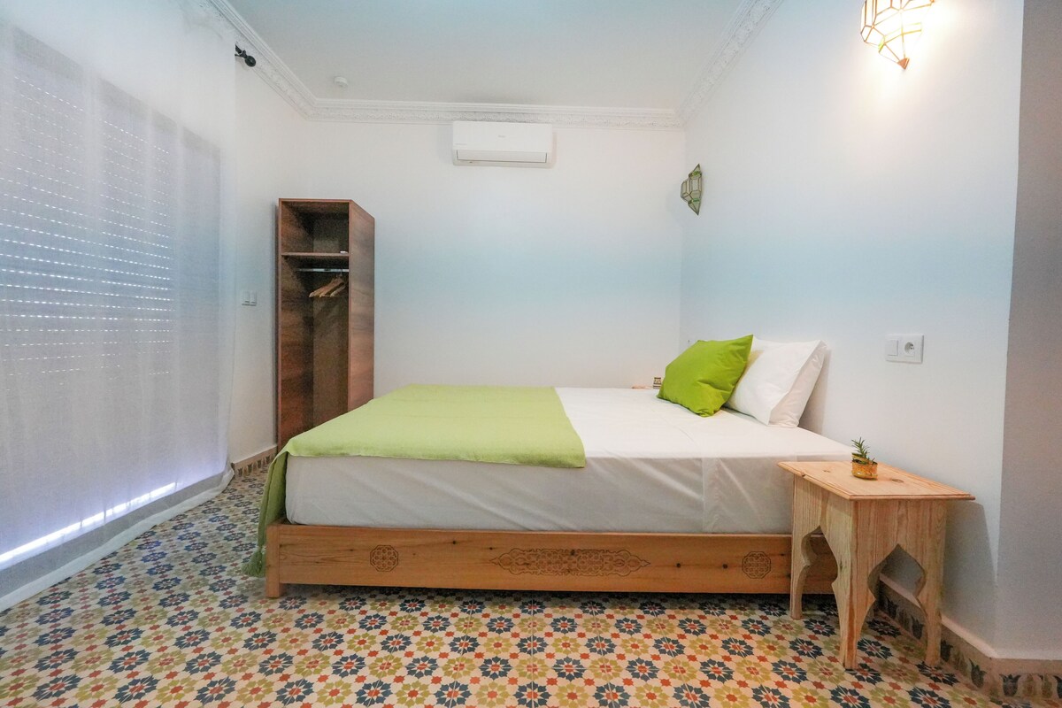 Un dormitorio con una cama, una mesita de noche de madera, un almohadón verde, un aire acondicionado instalado en la pared y un piso con estampado.