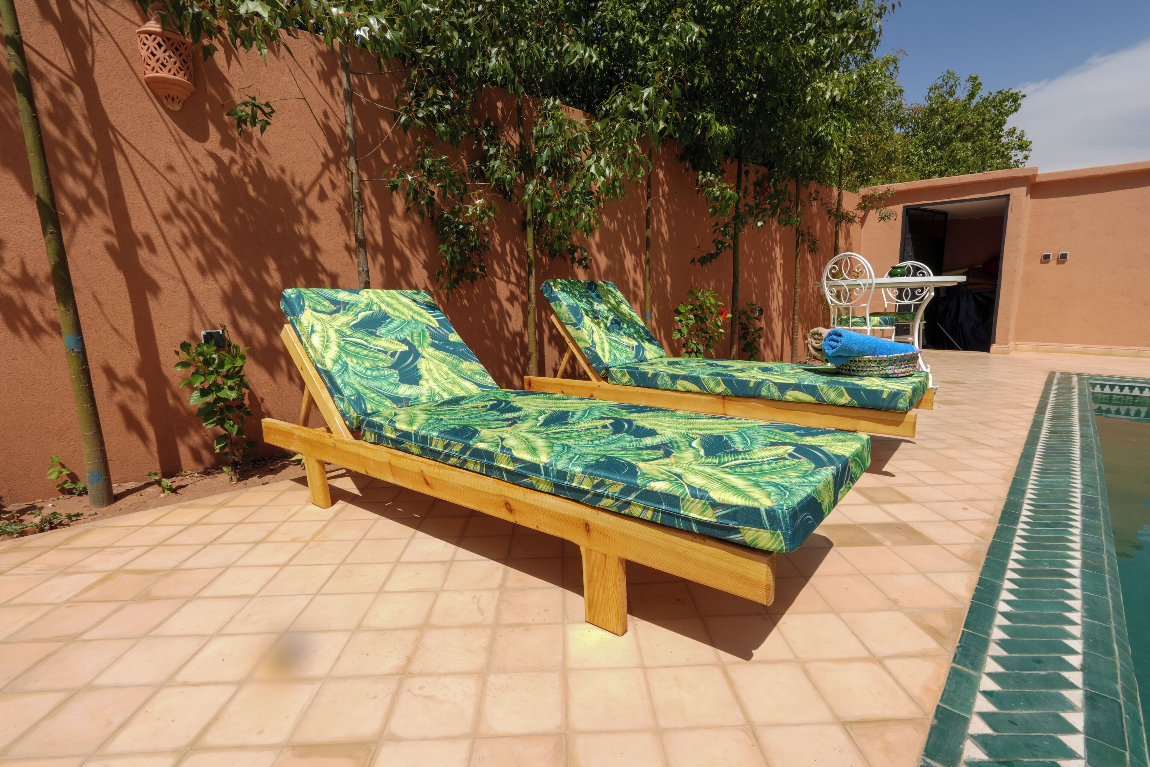 Deux fauteuils de jardin en bois avec des coussins à motifs, une terrasse carrelée et un espace piscine.