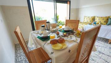 A dining table set with breakfast items, including croissants, eggs, and orange juice, in a room with a bed and a view of greenery.