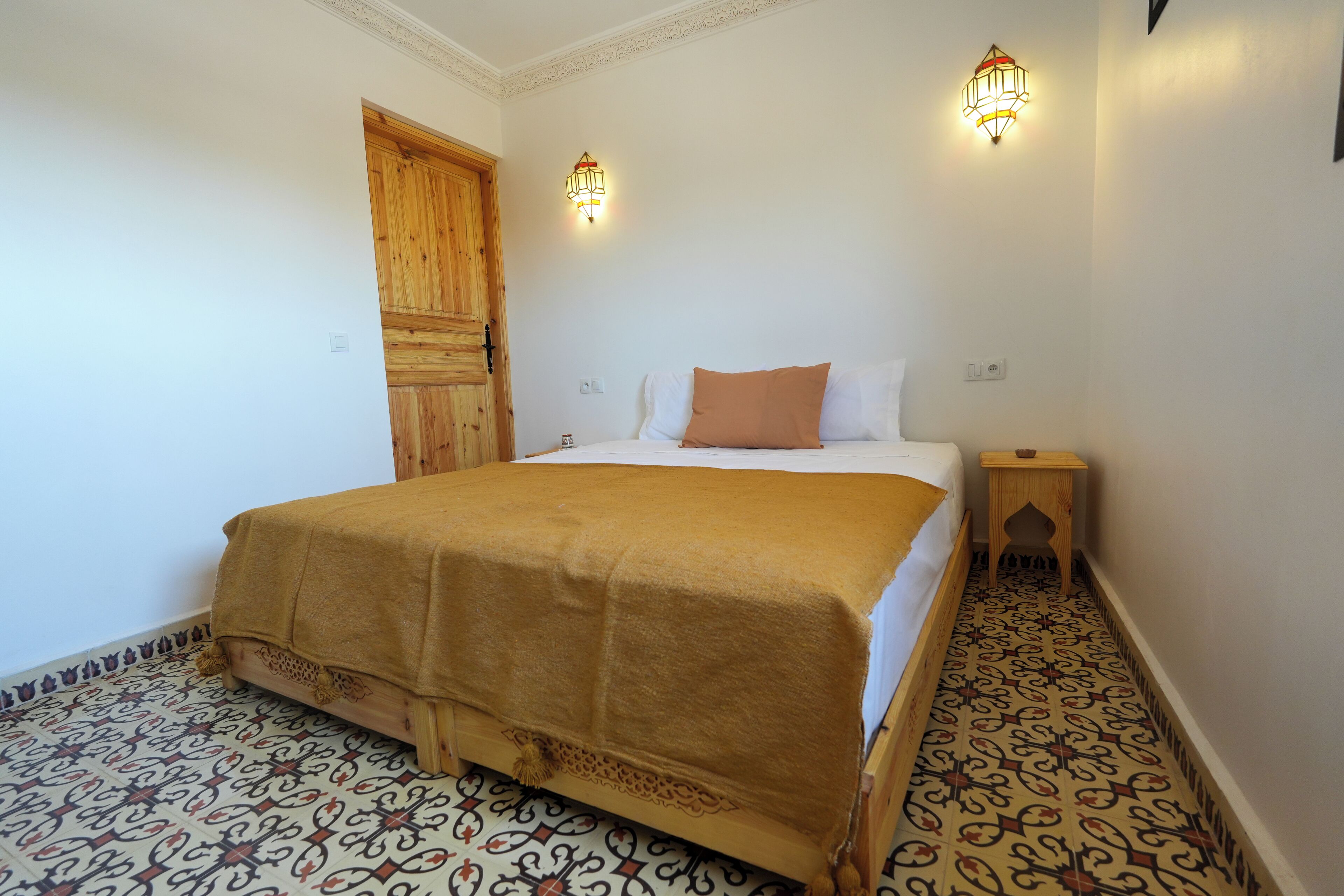 A bedroom with a bed, a wooden door, a nightstand, and patterned floor tiles.