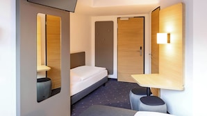 Habitación de hotel pequeña con una cama individual, una mesita de noche, un taburete, un espejo y un televisor montado en la pared.