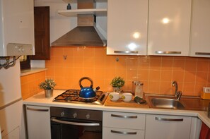 Una cocina con gabinetes blancos, un salpicadero de azulejos naranjas, una estufa a gas con un hervidor azul y un lavamanos de acero inoxidable.
