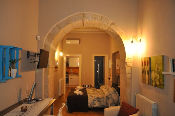 Una habitación acogedora con un arco de piedra que da a una cocina, una cama con ropa de cama floral y una mesa con lámpara.