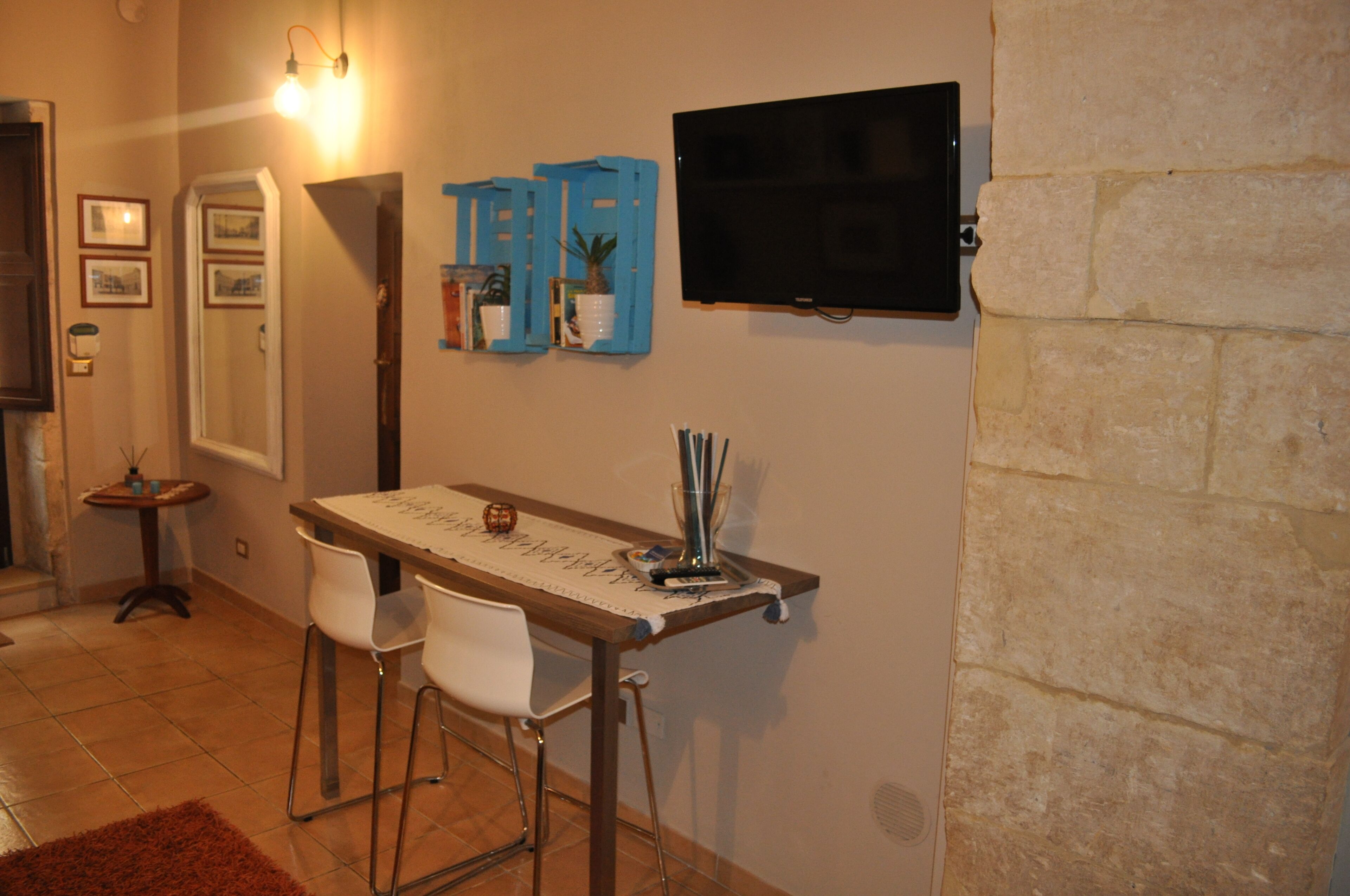 A dining area with a table, chairs, a TV mounted on the wall, and a small side table with a lamp.