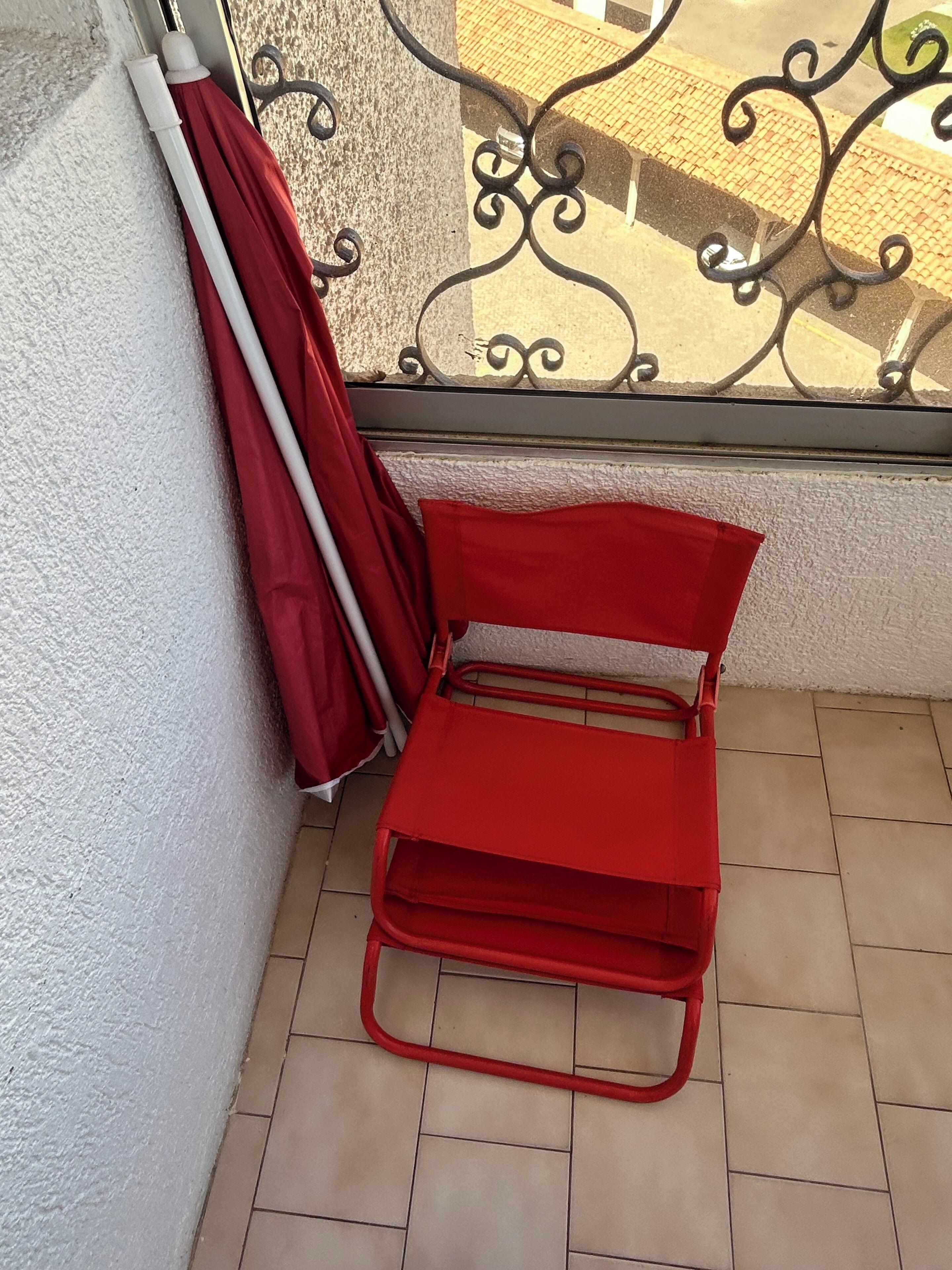 Una silla metálica roja con respaldo y una sombrilla roja apoyada contra una pared con textura.