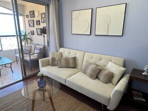 Een woonkamer met een beige bank, een glazen salontafel en twee abstracte schilderijen aan de muur.