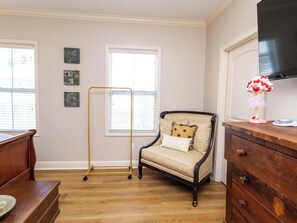 Een kamer met een houten vloer, een vintage fauteuil met gestreepte kussens, een houten dressoir, een goudkleurige standaard en een aan de muur bevestigde televisie.