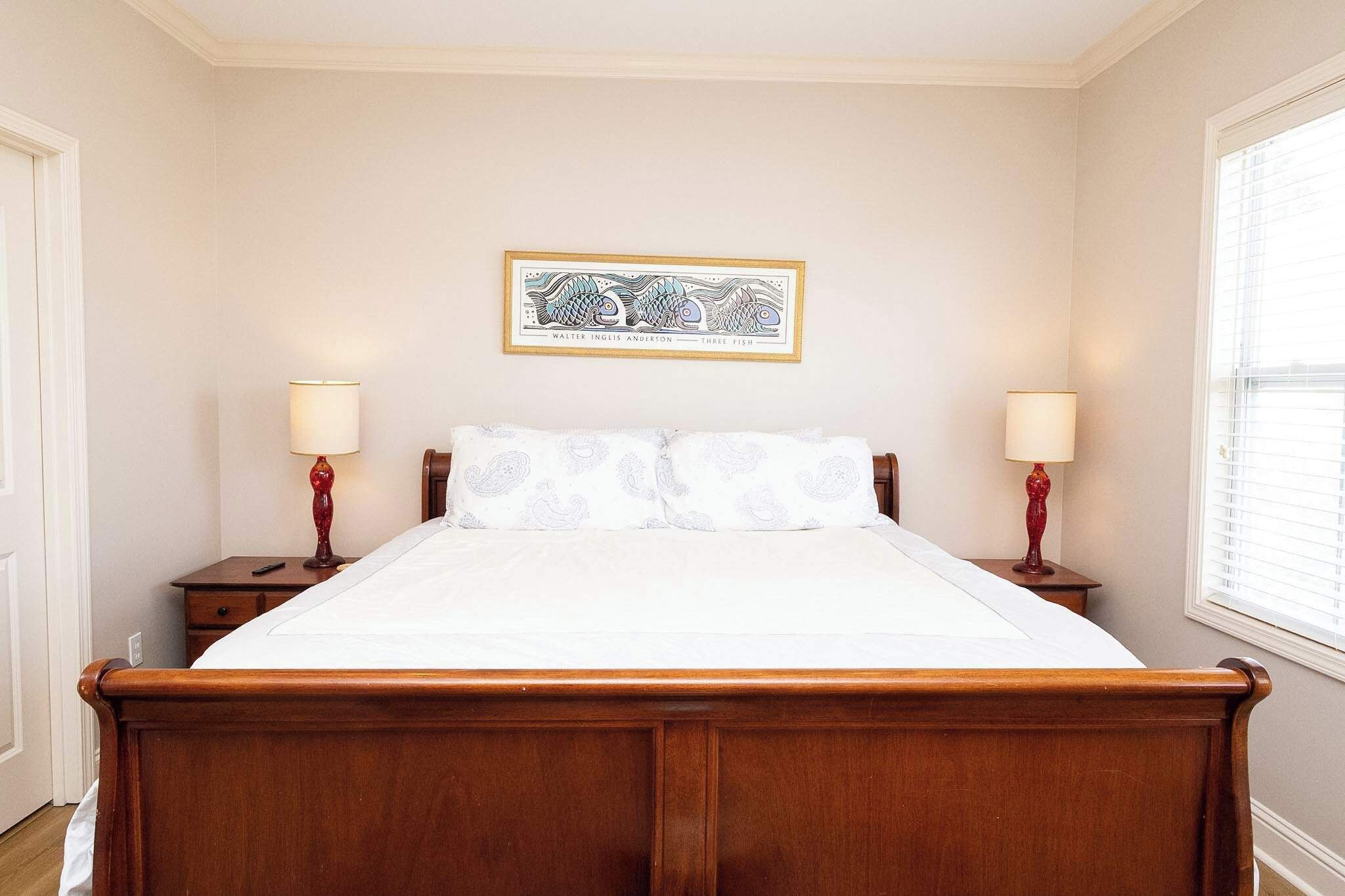 A bedroom with a wooden bed, two bedside lamps, a framed artwork on the wall, and a window with blinds.