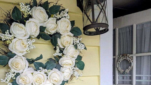 Una puerta amarilla con un arreglo floral blanco y un farol negro.
