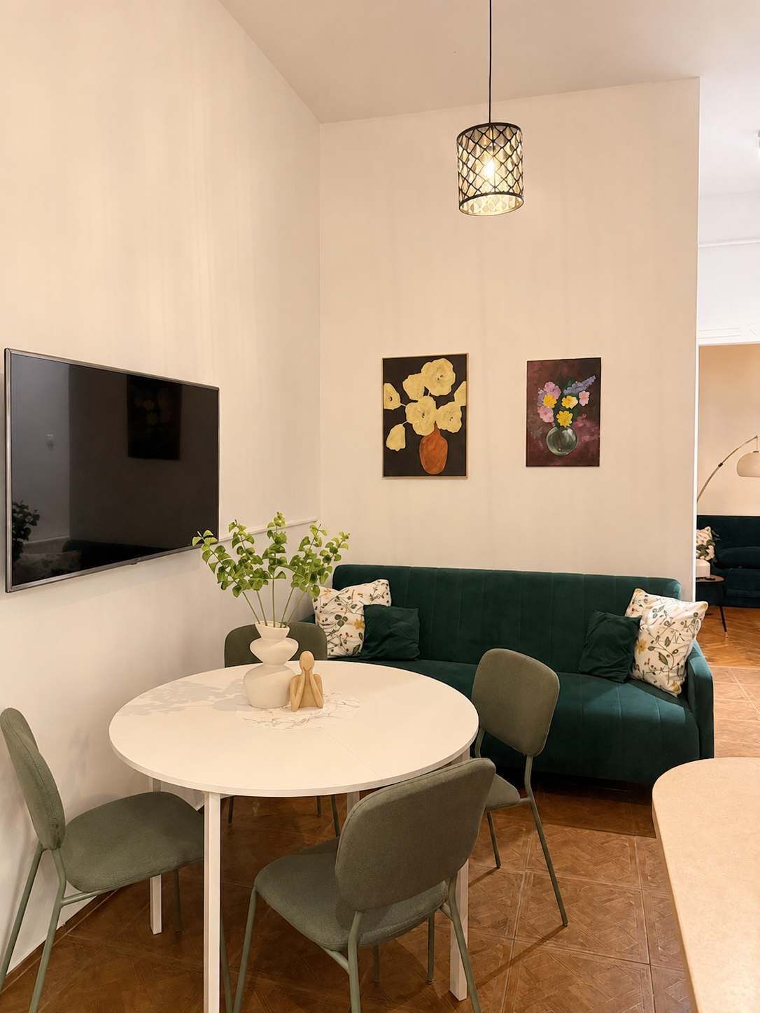 A modern living space with a round dining table, two green sofas, a flat-screen TV mounted on the wall, and two wall-mounted paintings.