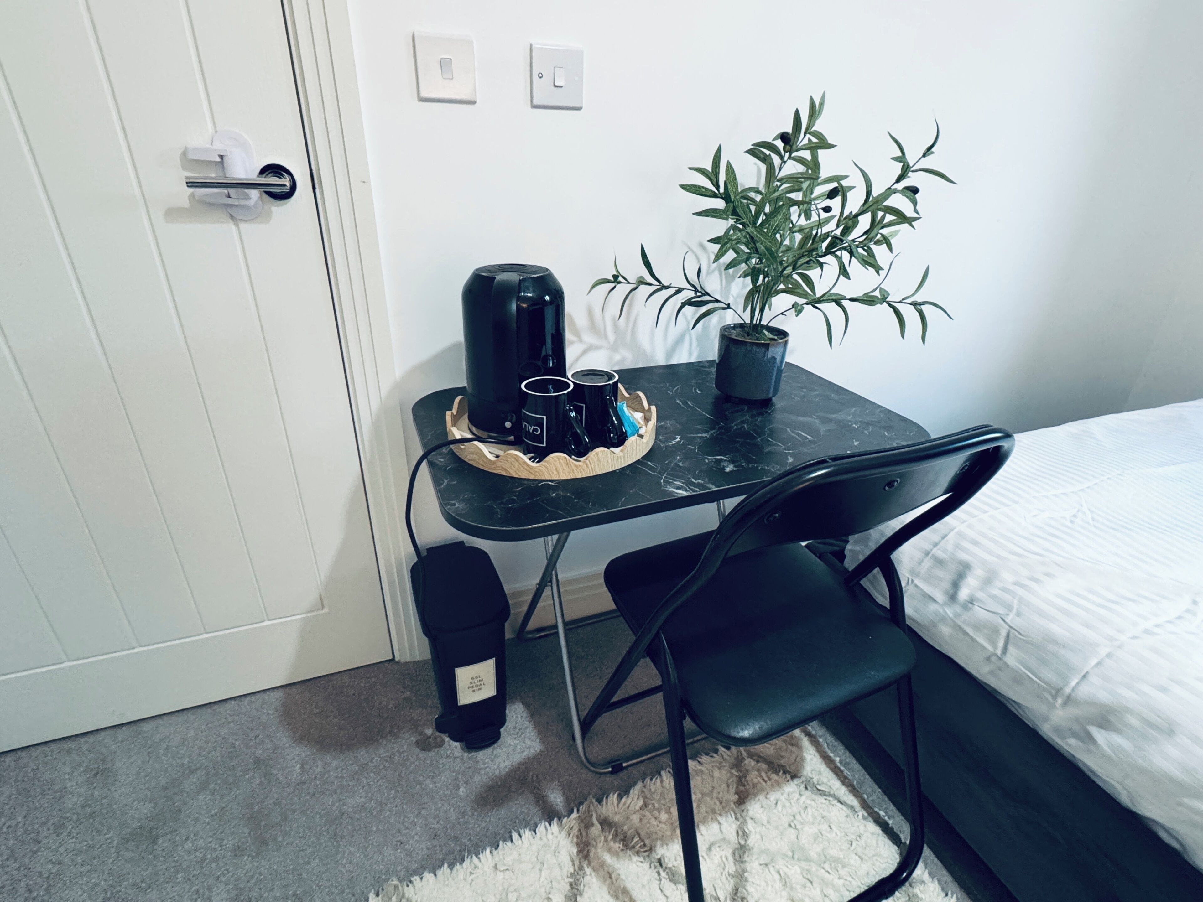 A small room with a bed, a desk with a coffee maker, a chair, a potted plant, and a trash bin.