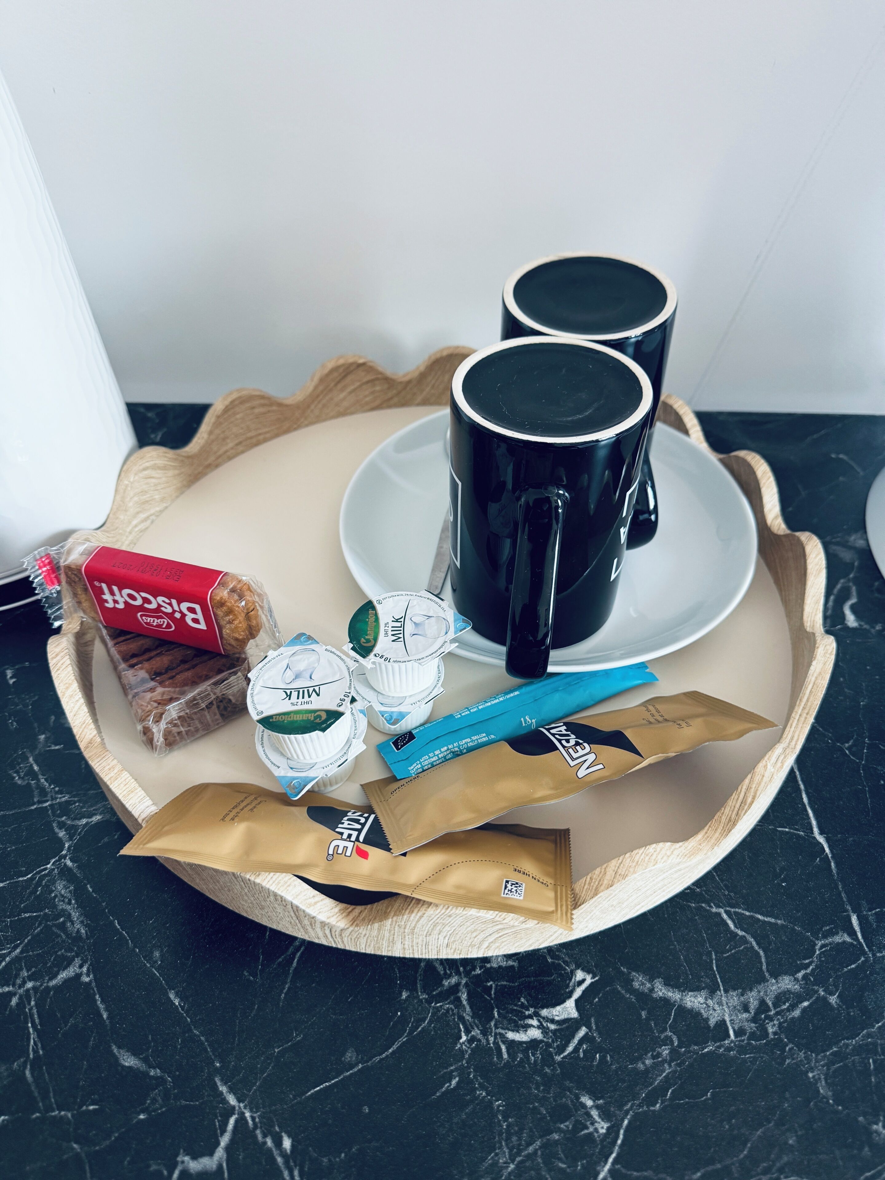A tray with two black mugs, a plate, a loaf of bread, a pack of biscuits, two small containers of milk, and two packets of coffee.