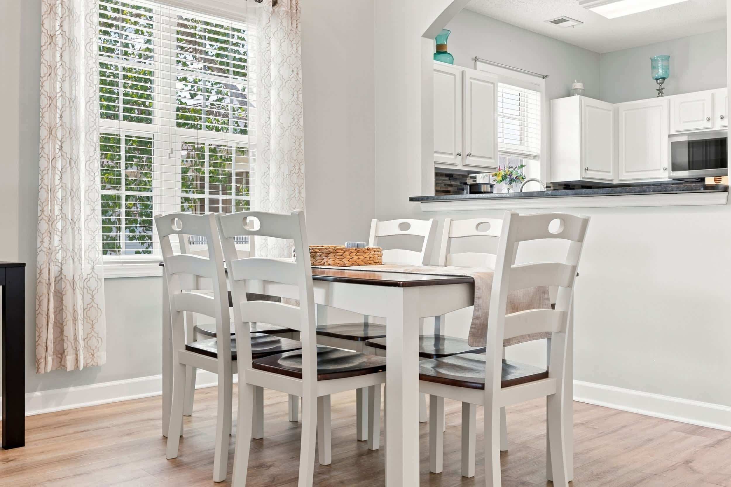 A dining area with a table and four chairs, a window with patterned curtains, and a kitchen in the background.