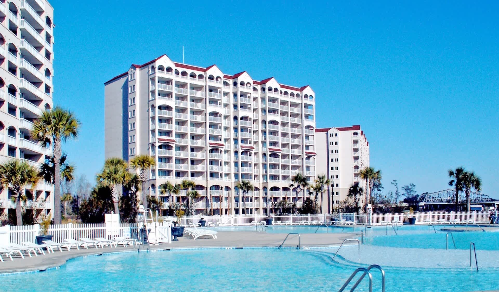 A hotel with multiple floors, a swimming pool, and palm trees.