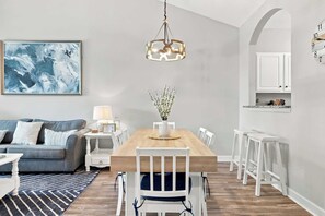 Ein modernes Wohnzimmer mit Essbereich, ausgestattet mit einem Holztisch, weißen Stühlen, einem blauen Sofa, einem Teppich und einem Gemälde an der Wand.