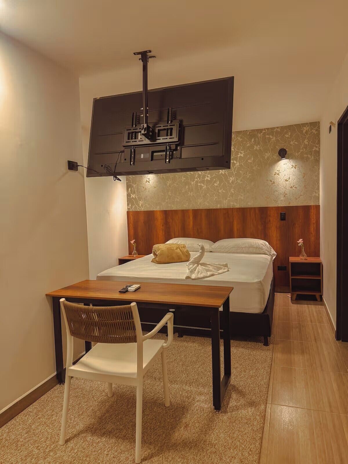 Una habitación de hotel con una cama, mesita de noche, una silla y un televisor montado en la pared.