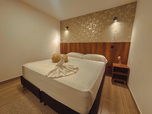 Una habitación de hotel con una cama grande, un cabecero de madera, una mesita de noche y una lámpara fijada a la pared.