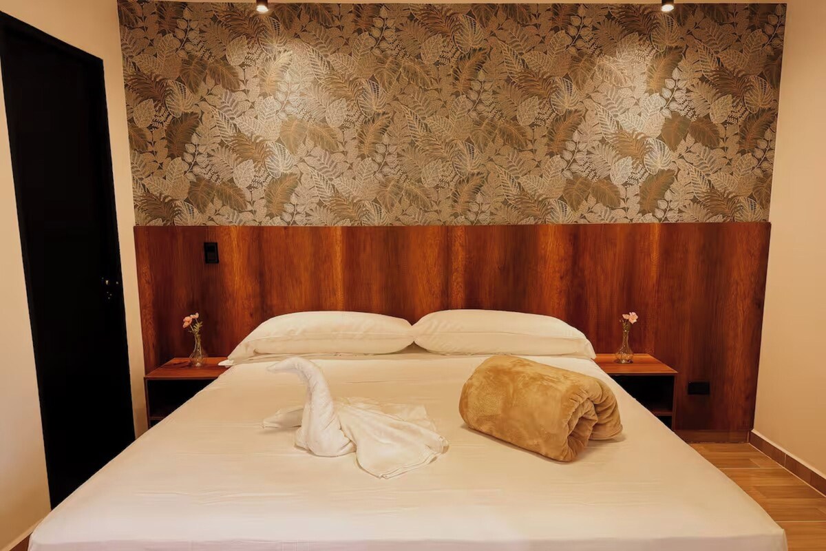 Habitación de hotel con una cama grande, cabecera de madera, dos mesitas de noche con lámparas y un papel tapiz estampado con motivos florales.