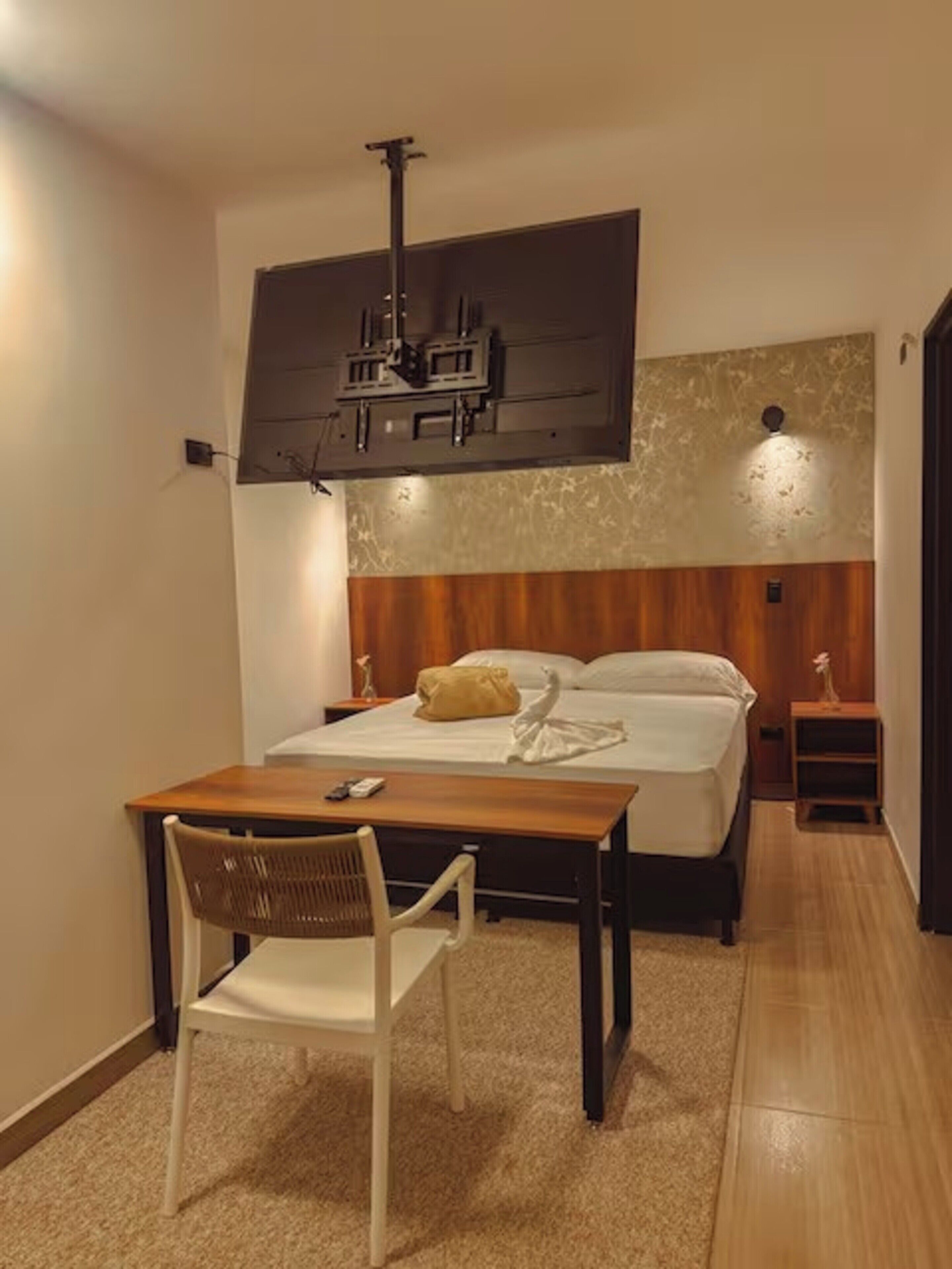 Habitación de hotel con cama, escritorio con silla, una mesita, televisor montado en la pared y lámpara.