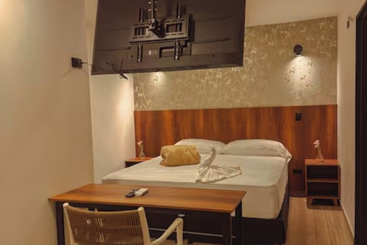 Habitación de hotel con cama, escritorio con silla, una mesita, televisor montado en la pared y lámpara.