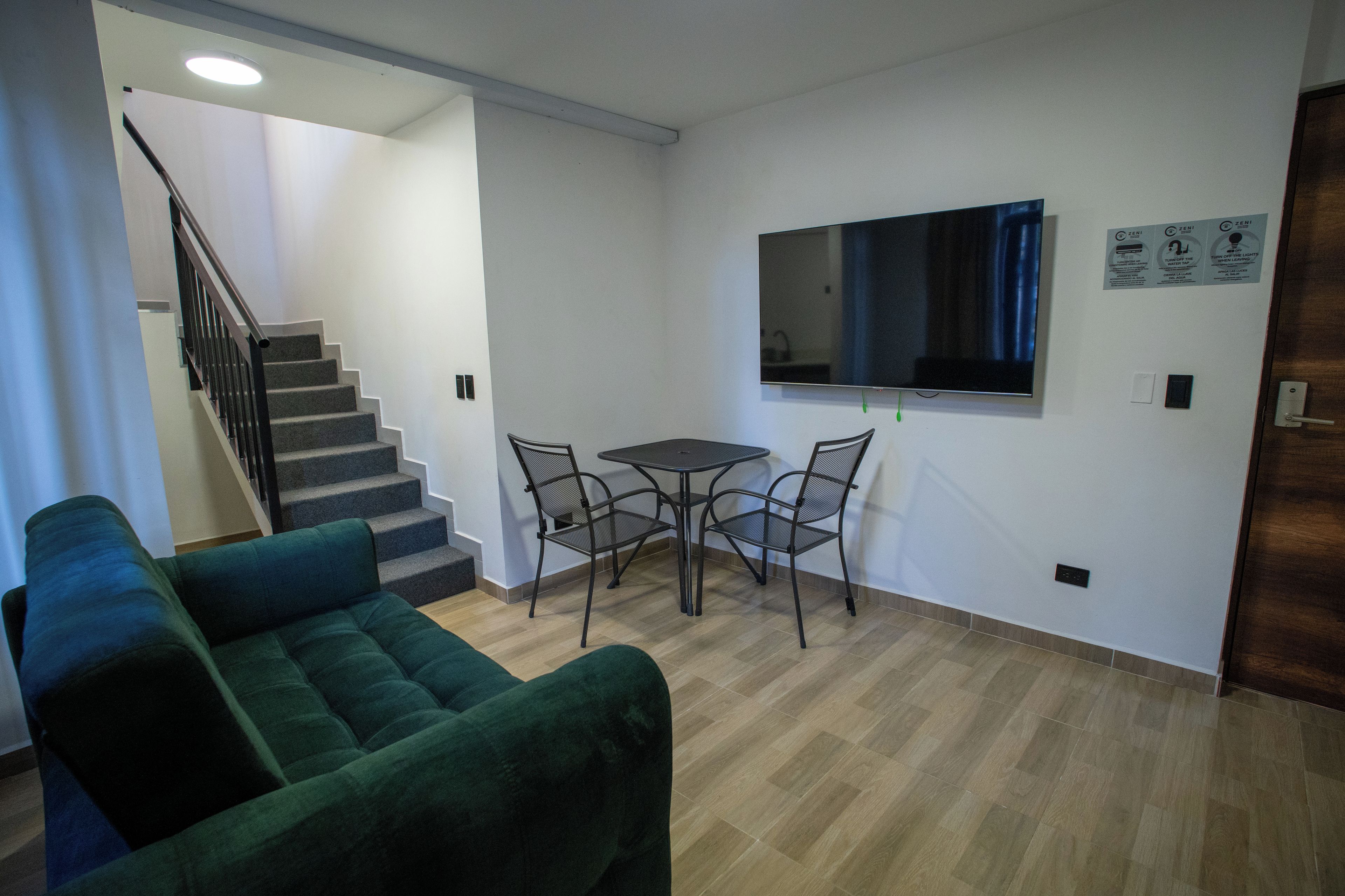 Un salon avec un canapé vert, une petite table entourée de chaises, un téléviseur à écran plat fixé au mur et un escalier menant à un autre niveau.
