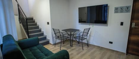 A living room with a green sofa, a small table with chairs, a flat-screen TV mounted on the wall, and a staircase leading to another level.