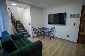 A living room with a green sofa, a small table with chairs, a flat-screen TV mounted on the wall, and a staircase leading to another level.