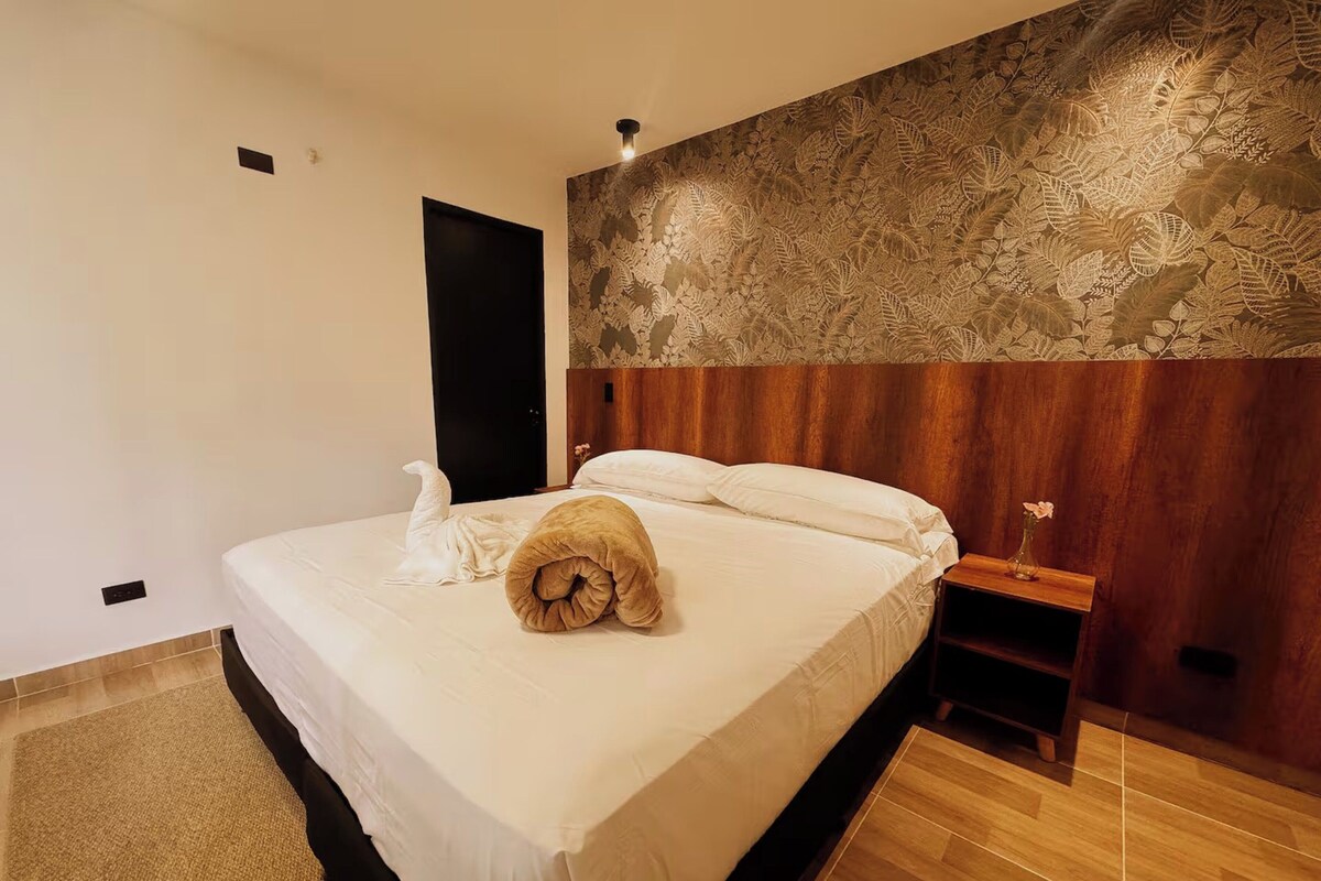 Habitació d'hotel amb llit, fusta de capçal, taula de nit i paret amb dibuixos de fulles.