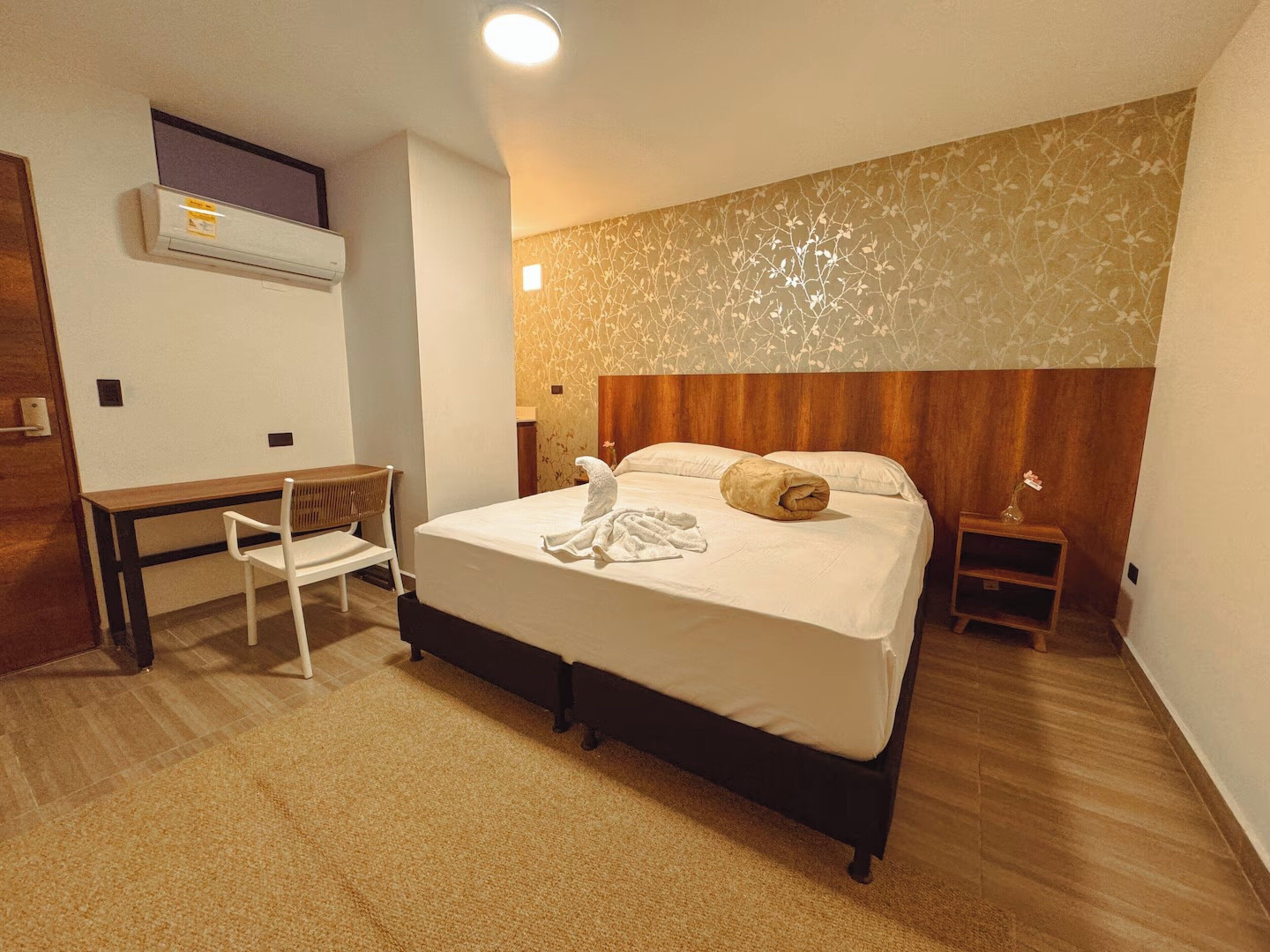 Ett hotellrum med en säng, ett skrivbord, en stol, ett nattduksbord och en väggmonterad luftkonditioneringsanl äggning.