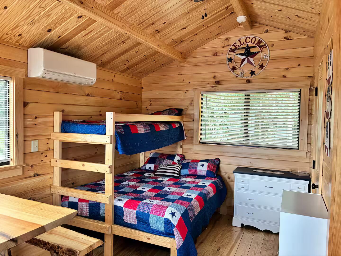 A cabin interior with bunk beds, a desk, a dresser, and a window with blinds.