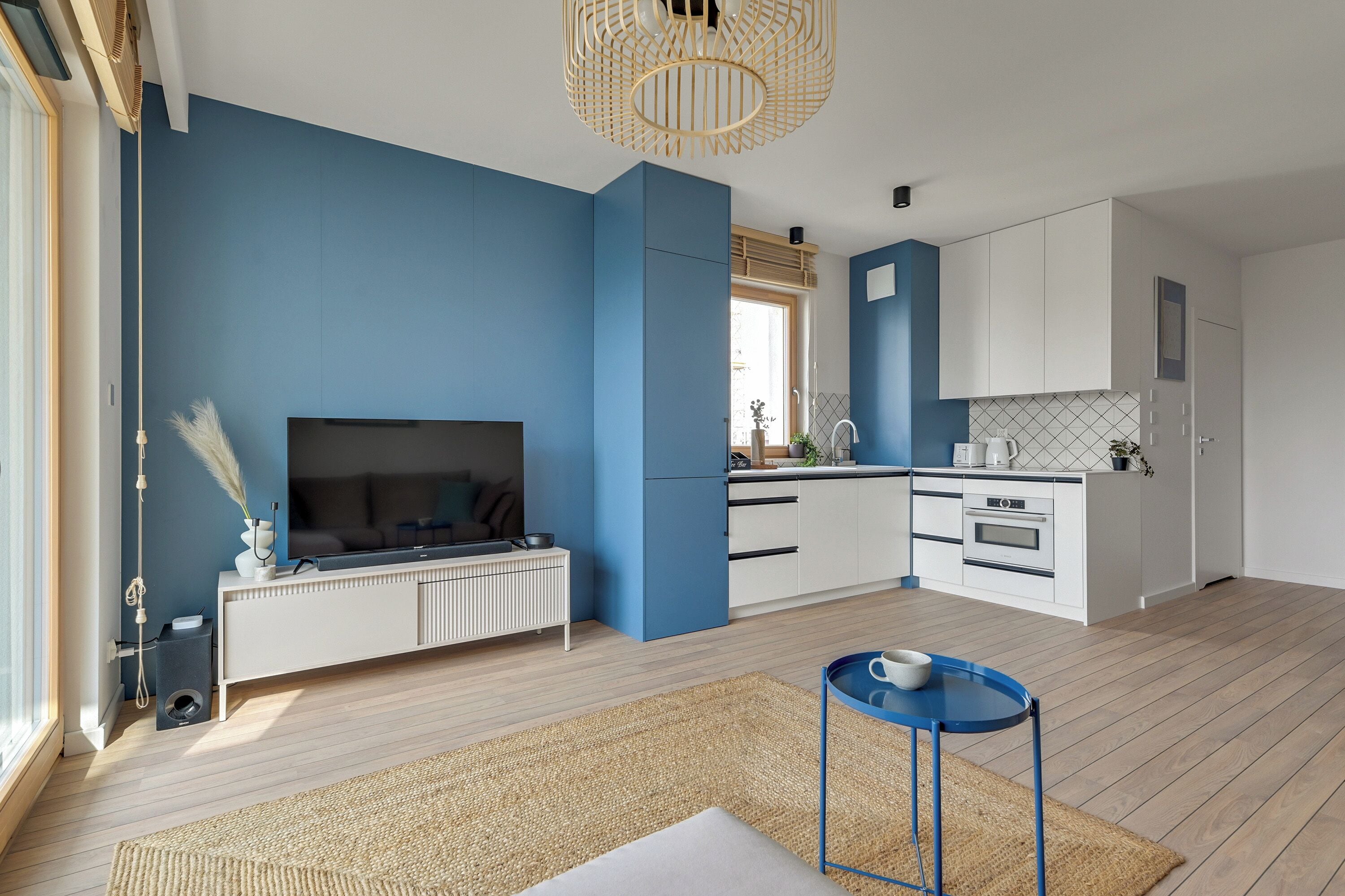 A modern kitchen with a blue accent wall, a dining area with a blue table, and a living room with a flat-screen TV.