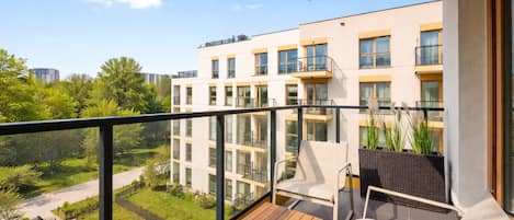 A balcony with a wooden table and chairs, overlooking a garden and a building with multiple windows.