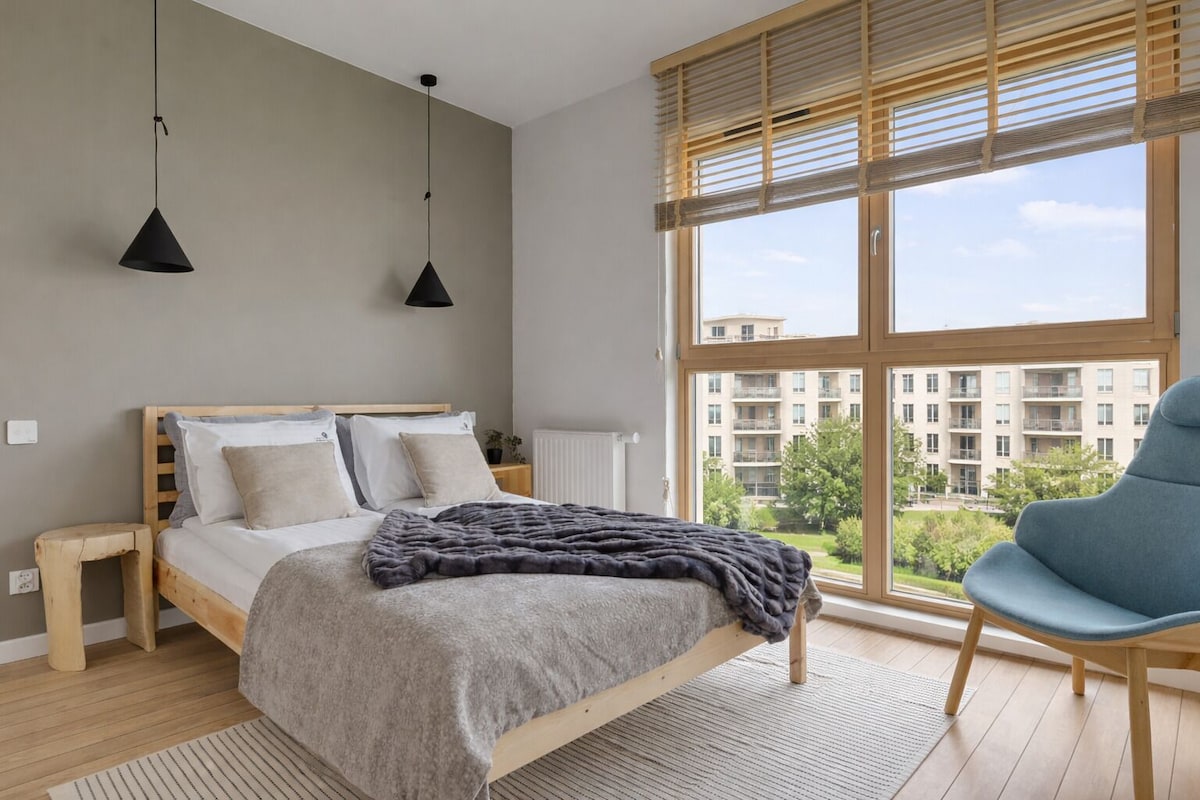 A modern bedroom with a large window, a bed with a grey blanket, wooden headboard, and two pendant lights.