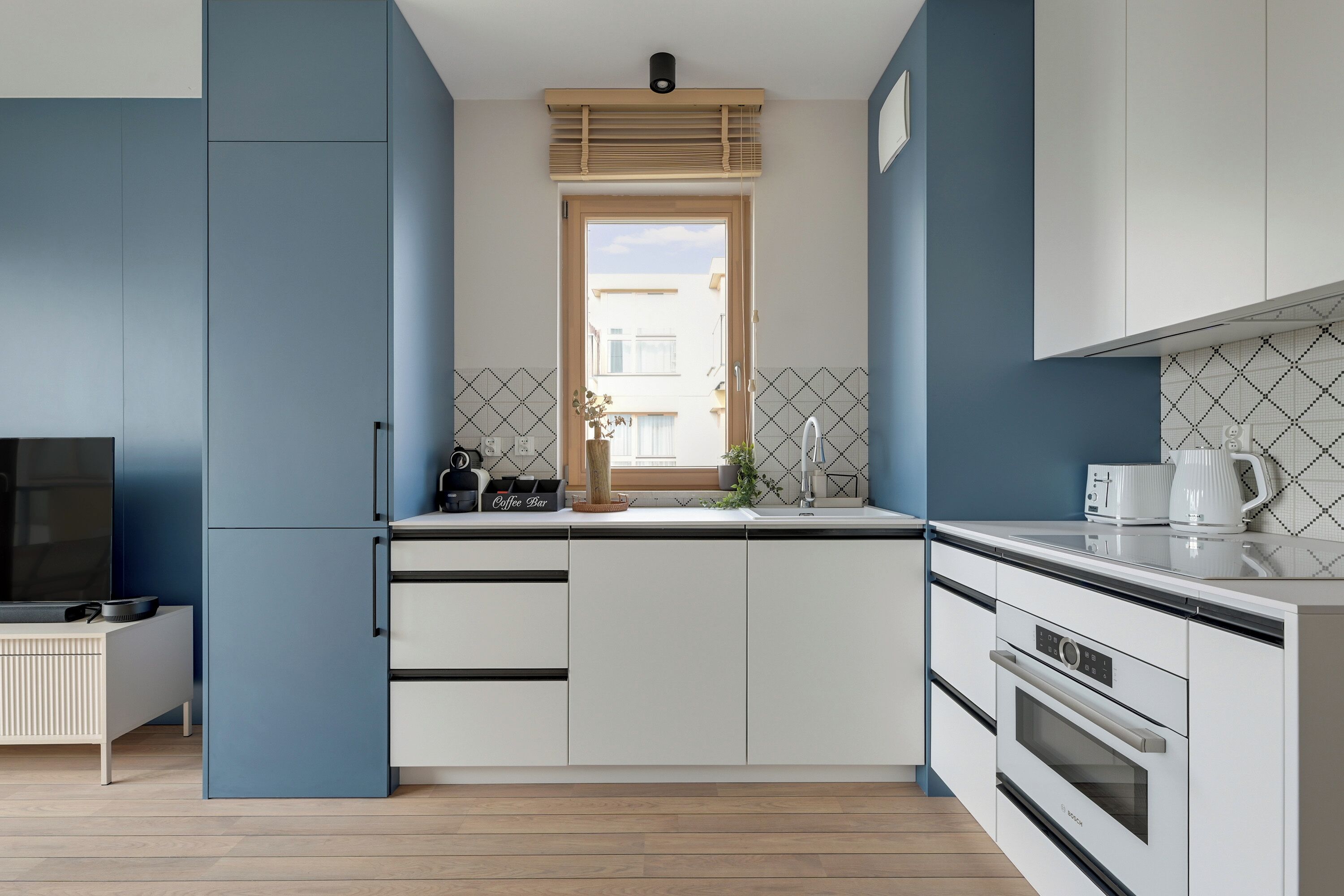 Cuina compacta amb armaris blancs, forn Bosch, cafetera i finestra amb vista a edificis.