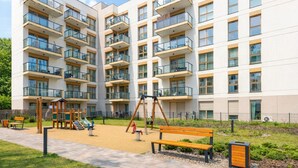 Un immeuble résidentiel à plusieurs étages doté de balcons, d’une aire de jeux avec des balançoires et des toboggans, et d’un chemin piétonnier pavé.