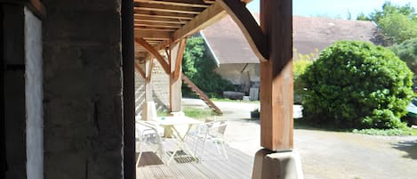 Una terraza de madera con mobiliario exterior, un muro de piedra y un espacio cubierto con vista a una casa y vegetación.