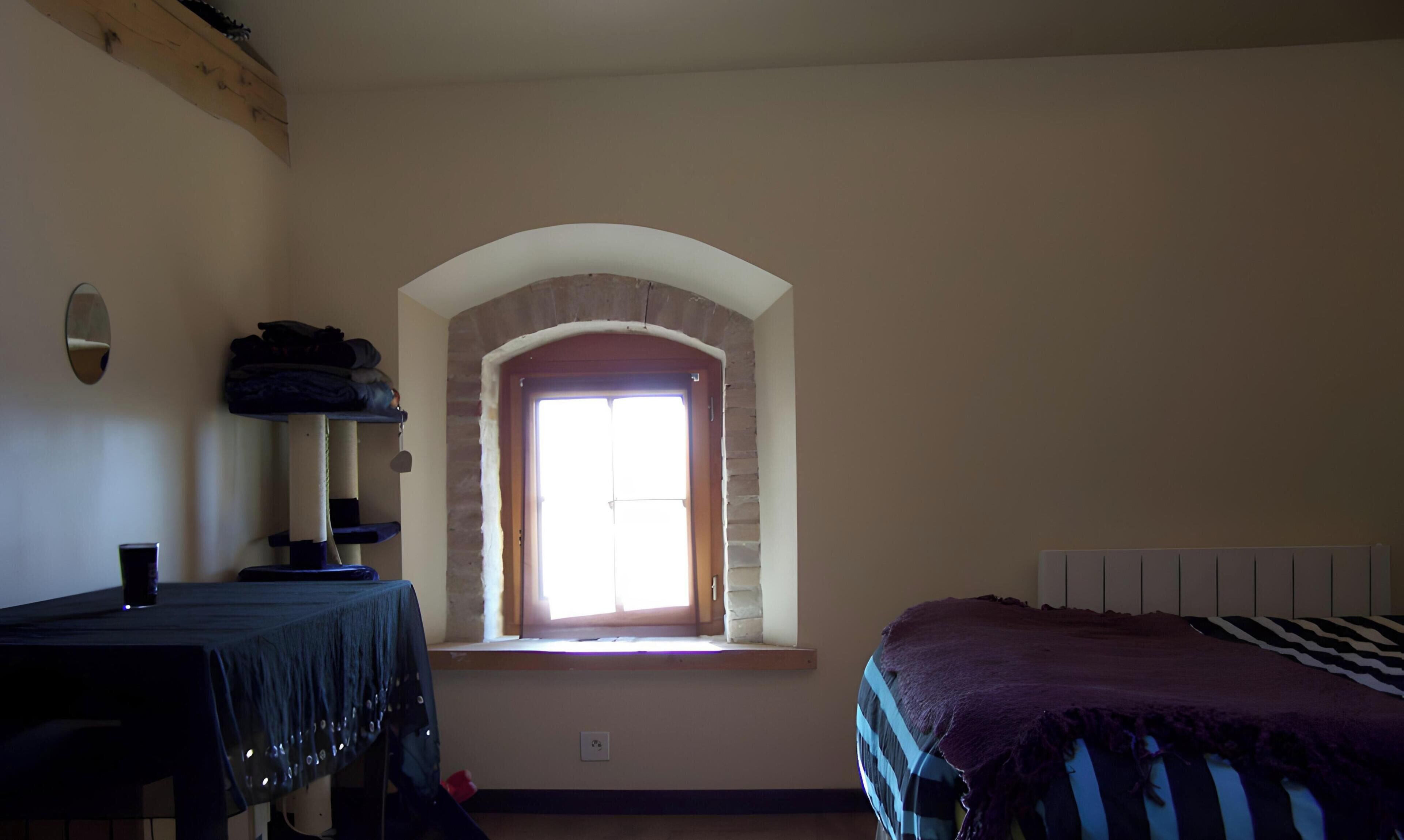 A room with a bed, a table, a window with a view, and a shelf with folded clothes.