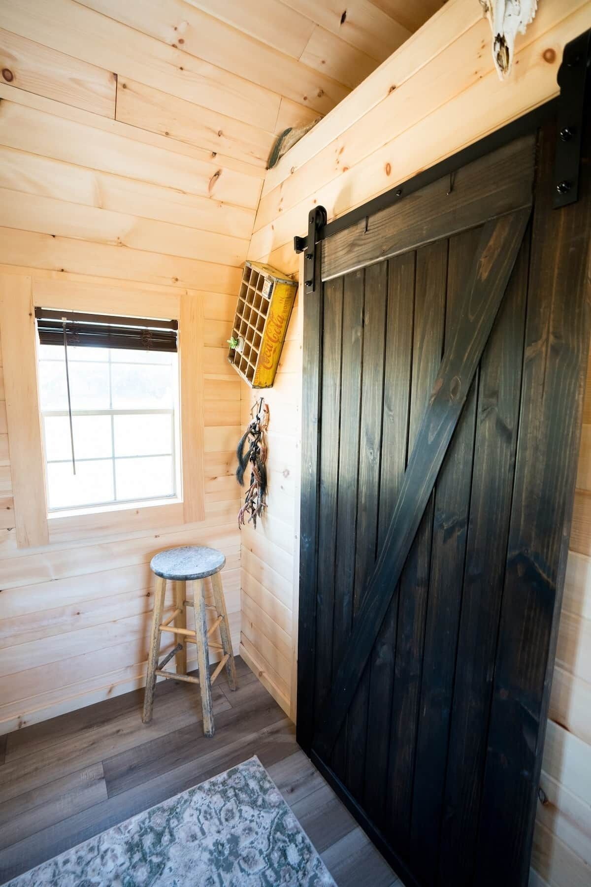Interior de una cabaña de madera con una puerta corredera grande, una ventana con persianas, un taburete pequeño y un fardel decorativo de Coca-Cola.