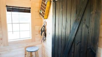 Interior de una cabaña de madera con una puerta corredera grande, una ventana con persianas, un taburete pequeño y un fardel decorativo de Coca-Cola.