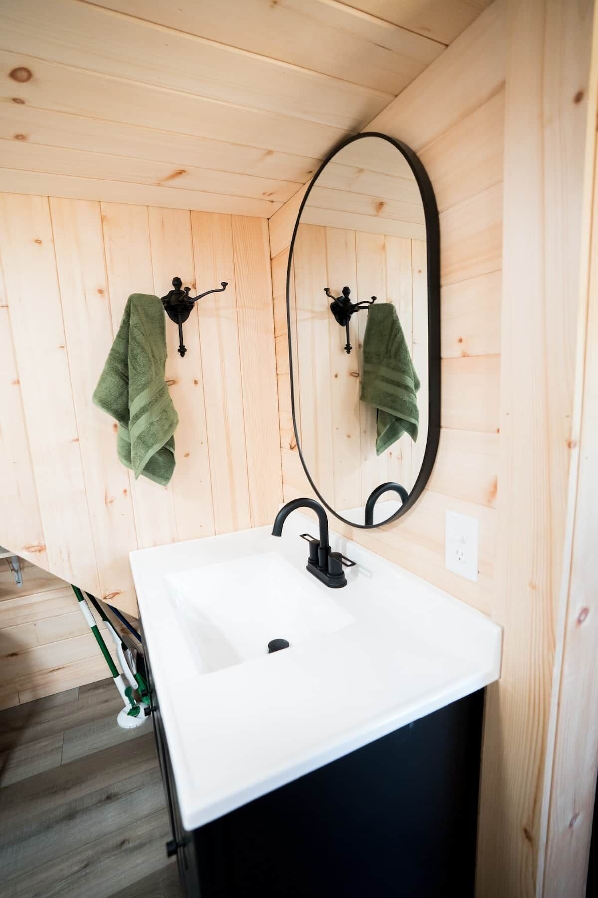 A bathroom with a white sink, black faucet, and a large oval mirror. There are two green towels hanging on black towel hooks.