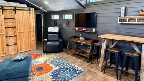 A room with a wooden ceiling, a black leather chair, a wooden table with stools, and a colorful rug.