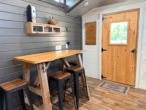 A dining area with a wooden table and stools, a shelf with decorative items, and a door with a window view. - The Roost | Hot Tub, Ponds & Farm Views (Carlisle County)