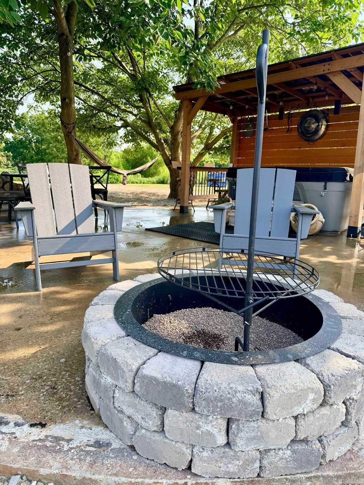 A fire pit with a metal grate, surrounded by stone. There are Adirondack chairs and a hammock in the background.