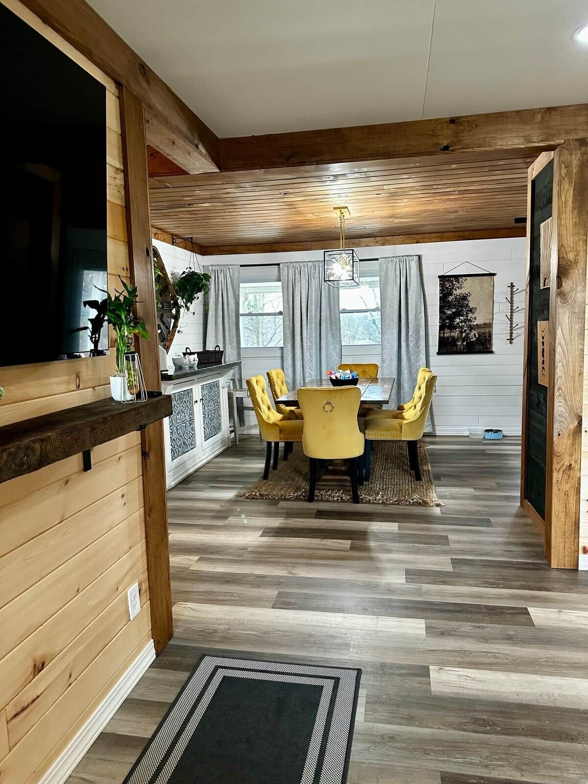 Un'area pranzo con soffitto in legno, un tavolo da pranzo con sedie gialle e un angolo cottura con una pianta e una finestra.
