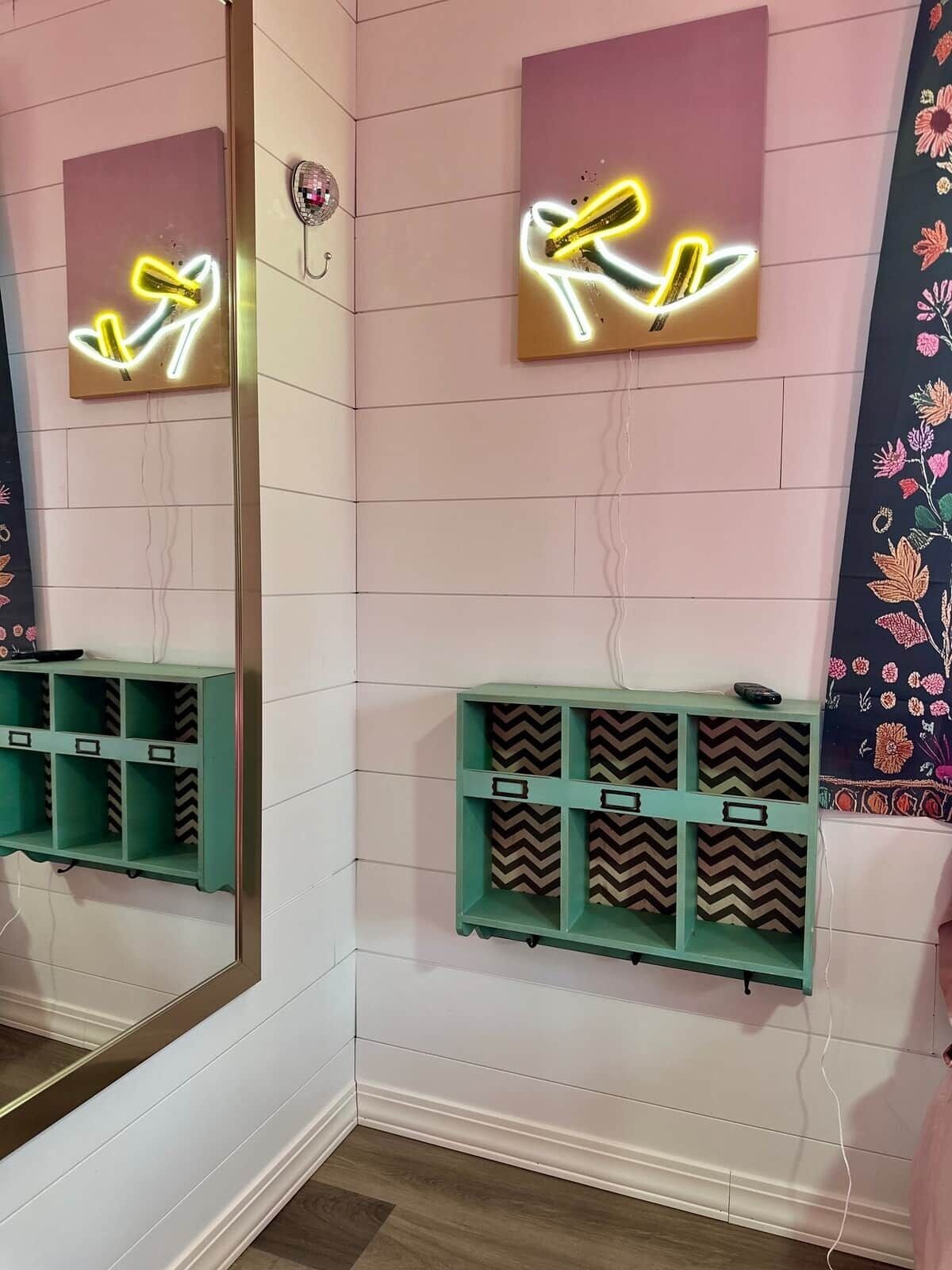 Une salle de bain avec un miroir, des œuvres d’art à base de néons et des étagères de rangement.