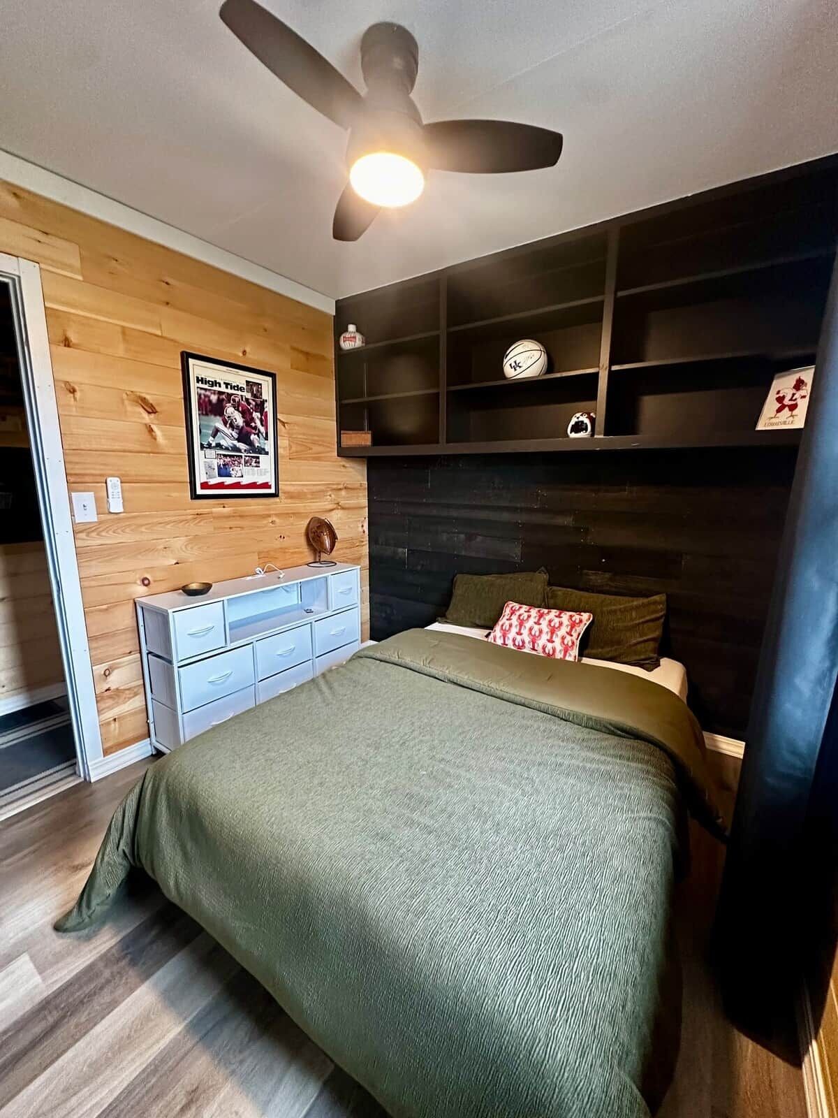 A bedroom with a bed, a ceiling fan, a wooden wall with a poster, a dresser, and a shelf with decorative items.
