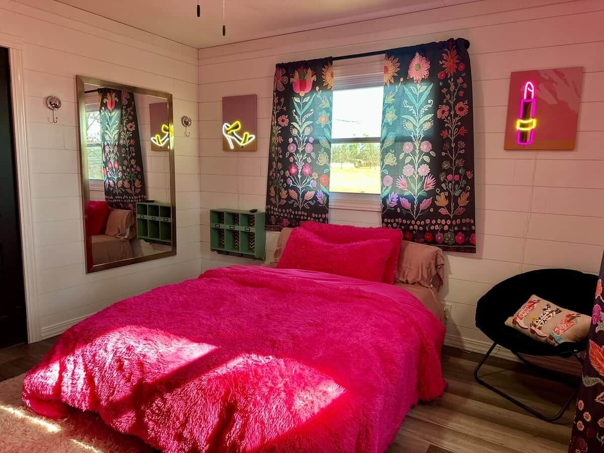 A bedroom with a large bed, a mirror, a window with floral curtains, and a neon lipstick sign.