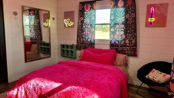 A bedroom with a large bed, a mirror, a window with floral curtains, and a neon lipstick sign.